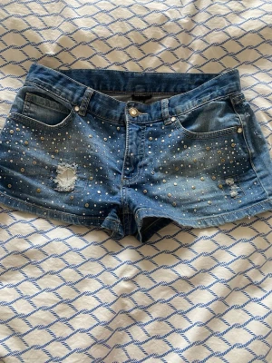 Blå jeansshorts med nitar och slitningar - Snygga blå jeansshorts dekorerade med silvriga nitar och pärlor framtill samt slitna detaljer. Klassisk femficksmodell med knapp och dragkedja. Perfekta för sommaren och ger en cool vibe till din outfit.