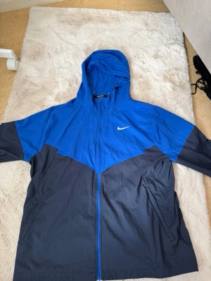 Blå Nike vindjacka med huva (Finns ej kvar i butik) - Snygg tvåfärgad vindjacka från Nike Running i blått och mörkblått. Jackan har huva, hel dragkedja framtill och sidofickor. Tillverkad i lätt, vindtätt material som passar perfekt för löpning eller utomhusaktiviteter. Nike-logga på bröstet.
