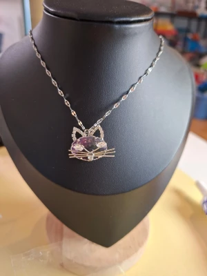 Halsband med kattansikte och kristaller - Snyggt halsband med ett hänge format som ett kattansikte. Hänget har glittrande stenar i silverfärg och lila, samt detaljer som öron och morrhår. Kedjan är silverfärgad och har ett tvinnat mönster. Perfekt för dig som gillar djur och unika smycken.