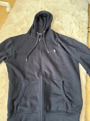 Marinblå hoodie från Polo Ralph Lauren - Snygg marinblå hoodie från Polo Ralph Lauren med dragkedja, två stora fickor framtill och klassisk huva med dragsko. Ikoniska lilla loggan broderad på bröstet. Tillverkad i mjukt bomullsmaterial som är skönt att ha på sig.