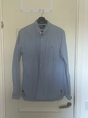 Blå skjorta från Polo Ralph Lauren - Klassisk ljusblå skjorta från Polo Ralph Lauren med button-down krage och broderad logga på bröstet. Skjortan har lång ärm, knappar framtill och är tillverkad i bomull. Perfekt för en clean och stilren look.