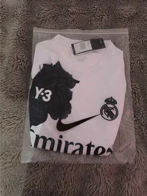 Vit Real Madrid Y-3 x Nike t-shirt - Vit helt ny och oanvänd t-shirt från Y-3 x Nike i samarbete med Real Madrid. Stor svart ros och Y-3-tryck på bröstet, Real Madrid-logga och Nike swoosh. Stort 'Emirates Fly Better'-tryck framtill. Tillverkad i lätt och skönt material. STORLEK XS/S