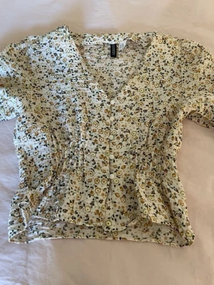 Blommig blus från H&M med knappar - Söt blommig blus från H&M med små blommor. Blusen har v-ringning, knappar framtill och korta ärmar. Smockad midja ger en fin passform och lätt volang nertill.  Denna är ifrån hm i storlek xxs