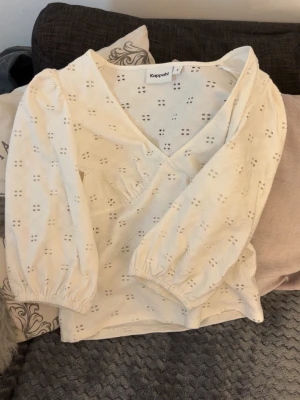 Vit topp  - Superfin vit blus/topp från Kappahl med broderade hålmönster och v-ringning. Blusen/toppen har puffiga långärmar med resår vid ärmslut och är gjord i ett lätt tyg som känns luftigt och fräscht. Perfekt att styla med jeans eller kjol. Oanvänd🩷