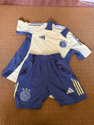 Ajax fotbollsträningsset  - Ajax träningsset från Adidas med gul t-shirt och mörkgrå shorts. T-shirten har svarta sidopaneler och shorts med Ajax-logga, tre stjärnor och gula detaljer. Perfekt för fotbollsträning eller gymmet. Materialet är lätt och andas.