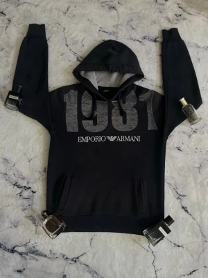 Svart hoodie Emporio Armani 1981 - Svart hoodie från Emporio Armani med stort tryck '1981' och logga på bröstet. Klassisk huva med dragsko och känguruficka framtill. Mjuk insida och ribbade muddar. Perfekt för en stilren och avslappnad look.