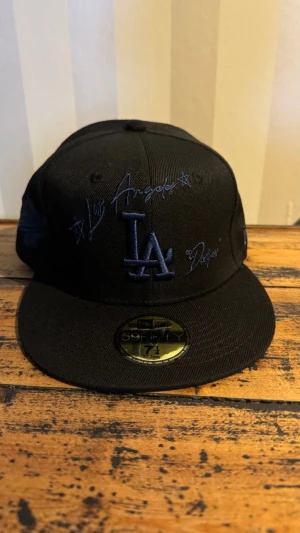 Svart LA Dodgers keps New Era - Snygg svart keps från New Era med LA Dodgers brodyr i mörkblått framtill och coola detaljer på sidan. Klassisk flat peak och broderade ventilationshål. Perfekt för dig som gillar streetwear och sportig stil.