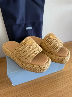 Prada Raffia sandaler - Snygga beige platåsandaler Prada med flätad design i raffia och bred rem över foten. Sandalerna har en chunky sula och ikonisk Prada-logga framtill. Perfekta för en trendig sommarlook och ger en cool vibe till outfiten.