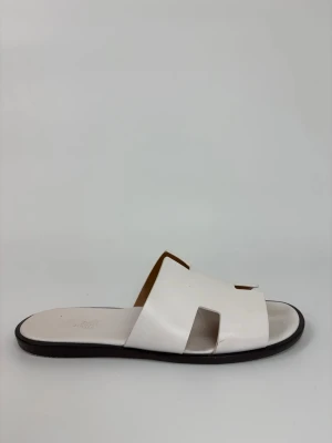 Vita slip-in Hernes sandaler i läder - Stilrena vita sandaler i äkta läder med breda remmar och öppen tå. Slip-in modell med svart sula och minimalistisk design, perfekta för en avslappnad och trendig look. Passar dig som gillar enkelhet och klassisk stil.