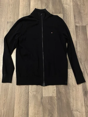 Mörk blå ziptröja från Tommy Hilfiger - Mörk blå långärmad ziptröja från Tommy Hilfiger med liten broderad logga på bröstet. Tröjan har hög krage och ribbade muddar. Perfekt för en stilren och enkel look.