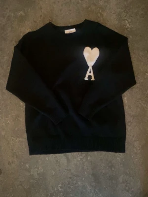 Svart stickad tröja från Ami Paris - Svart stickad tröja från Ami Paris med vitt hjärta och bokstaven A broderat på bröstet. Klassisk rund halsringning och långa ärmar. Perfekt för dig som gillar stilrena och trendiga plagg.