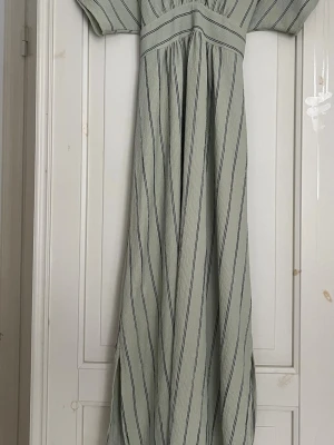 Randig långklänning- MNG Suit - Ljusgrön långklänning med smala svarta ränder och korta ärmar. Klänningen har ett markerat liv och ett brett knytband i midjan som ger en snygg siluett. Perfekt för dig som gillar en avslappnad men ändå stilren look. Klänningen är från Mango i storlek S. 