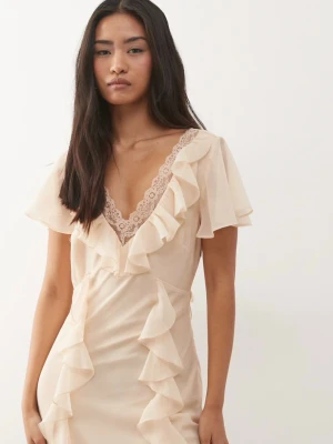 Beige långklänning med volang - Romantisk beige långklänning med v-ringning, spetsdetalj och volanger längs hela klänningen. Klänningen har korta ärmar och är tillverkad i ett lätt, flowy material som ger en elegant siluett. Perfekt till studentbalen eller midsommar!