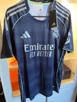 Real Madrid matchtröja Adidas blå L - Snygg blå Real Madrid matchtröja från Adidas med vita detaljer och klubbmärke på bröstet. Tröjan har slim fit-passform, korta ärmar och trycket 'Emirates Fly Better' på framsidan. Perfekt för fotbollsträning eller att bära till vardags.