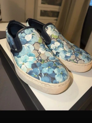 Gucci slip-on sneakers med blommor - Snygga slip-on sneakers från Gucci med blått och vitt blommönster på canvas och mörkblå detaljer. Skorna har tjock vit sula och klassisk GG-logga i bakgrunden. Perfekta för dig som vill sticka ut med exklusiv design och färgglad stil.