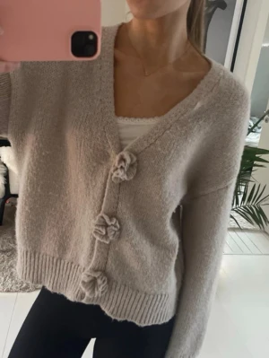 Beige kofta med blomknappar - Mysig beige kofta med v-ringning och tre dekorativa blomformade knappar framtill. Koftan är stickad i ett mjukt material och har långa ärmar samt ribbade muddar vid ärmslut och nederkant. Köpt från Nelly men märket är pieces.