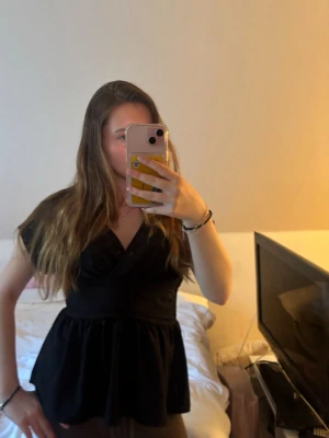 Svart omlottopp med v-ringning - Snygg svart omlottopp med v-ringning och korta ärmar. Toppen har en markerad midja och ett lätt utställd nederdel som ger en feminin siluett. Perfekt att styla med jeans eller kjol för en trendig look.