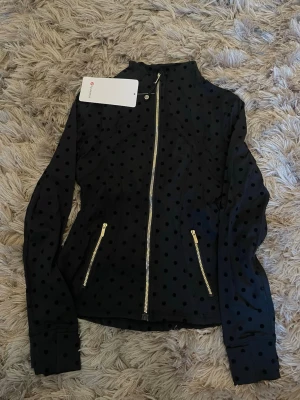 Svart prickig Lululemon jacka ny modell  - Snygg svart jacka med prickigt mönster och guldfärgade dragkedjor framtill och vid fickorna. Jackan har hög krage och lång ärm, perfekt för dig som vill ha en unik och trendig look. Materialet känns mjukt och följsamt.