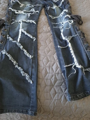 Svarta jeans med snörning och slitningar - Unika svarta jeans med kraftiga slitningar och fransade detaljer över hela benen. Sidorna har bred snörning med svarta band som ger en edgy och alternativ look. Perfekta för dig som gillar streetstyle och vill sticka ut. I storlek W31/L32