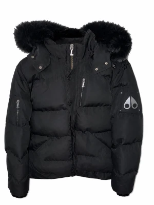 Svart dunjacka från Moose Knuckles - Säljer en svart dunjacka från Moose Knuckles med avtagbar huva och fuskpäls. Jackan har flera dragkedjeförsedda fickor, stora knappar och logotyp på ärmen. Perfekt för kalla vinterdagar tack vare tjockt dunfoder och vindtätt material.