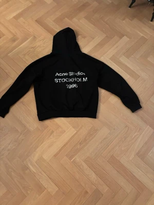 Svart hoodie Acne Studios Stockholm - Svart hoodie från Acne Studios med vit text på ryggen: 'Acne Studios STOCKHOLM 1996'. Klassisk passform med huva och ribbade muddar. Perfekt för dig som gillar stilrena och trendiga plagg med streetkänsla.