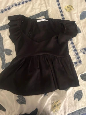 Svart volangtop från H&M - Snygg svart topp från H&M med volangdetaljer vid axlarna och en lätt utsvängd peplum nedtill. Toppen har rund halsringning och är tillverkad i ett mjukt tyg som ger en skön känsla. Perfekt för dig som gillar en feminin och trendig stil.