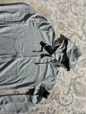 Grå hoodie från Stone Island - Snygg grå hoodie från Stone Island med dragkedja framtill och klassisk huva med dragsko. Ikonisk svart patch med gul och grön kompasslogga på ärmen. Tillverkad i mjukt bomullsmaterial och har två fickor framtill. Perfekt för en avslappnad och trendig stil.