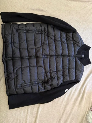Svart pufferjacka  från Moncler - Svart dunjacka från Moncler med quiltad framsida och stickade svarta ärmar och axelpartier. Jackan har dragkedja framtill och Moncler-logga på axeln. Perfekt för dig som gillar stilren och sportig look.