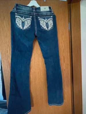 Mörkblå jeans med broderade fickor - Snygga mörkblå jeans från Rock Revival med raka ben och unika bakfickor dekorerade med broderade vingar och paljettdetaljer. Jeansen har kontrastsömmar och klassisk femficksdesign. Perfekta för dig som vill sticka ut med coola detaljer.