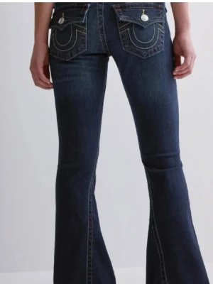 Blå bootcut jeans från True Religion - Säljer ett par mörkblå bootcut jeans från True Religion med ikoniska bakfickor och kontrastsömmar. Jeansen har låg midja och klassisk femficksdesign. Perfekta för dig som gillar en avslappnad men ändå trendig look.