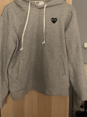 Grå hoodie från Comme des Garçons Play - Snygg grå hoodie från Comme des Garçons Play med svart hjärta och ögon-logga på bröstet. Hoodien har huva med vita snören och två fickor framtill. Perfekt för en chill och stilren look.