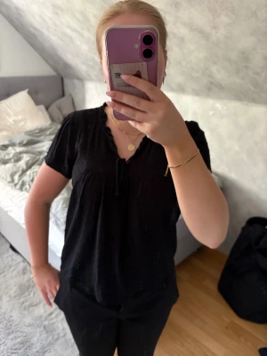 Svart blus med knyt och volang - Svart blus med korta ärmar, volangkant vid halsringningen och knytband framtill. Lätt och luftig modell med diskret struktur i tyget, perfekt för en avslappnad men ändå stilren look.