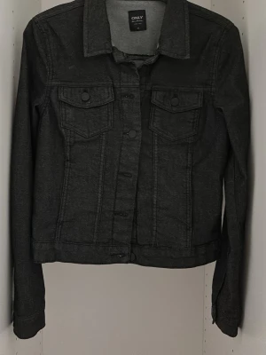 Svart jeansjacka från ONLY - Stilren svart jeansjacka från ONLY med klassisk krage, knäppning framtill och två bröstfickor med lock och knapp. Jackan har en figurnära passform och långa ärmar. Perfekt för dig som gillar en enkel men cool look.