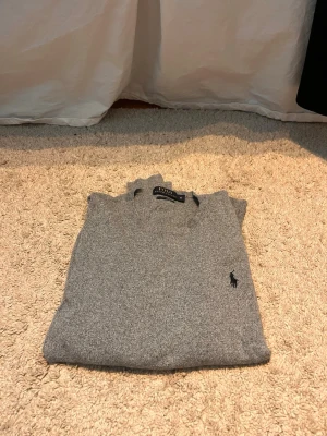 Grå tröja från Ralph Lauren - Klassisk grå tröja från Ralph Lauren med rund halsringning och diskret broderad logga på bröstet. Tröjan har lång ärm och är tillverkad i ett mjukt, tunt material som passar perfekt till lager på lager. Enkel och stilren design. Storlek M