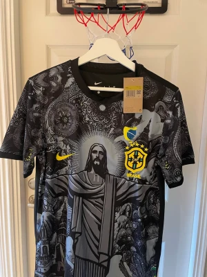 Nike Brasil svart fotbollströja - Snygg svart fotbollströja från Nike med Brasil-logga och gult tryck. Tröjan har ett detaljerat grafiskt mönster med Kristusstatyn och andra motiv, samt rund hals och korta ärmar. Perfekt för dig som vill sticka ut på planen eller heja på Brasilien.