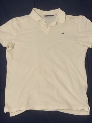 Vit pikétröja med krage Morris - Klassisk vit pikétröja med krage och korta ärmar. Liten broderad logga på bröstet. Perfekt till jeans eller shorts för en clean och enkel stil.