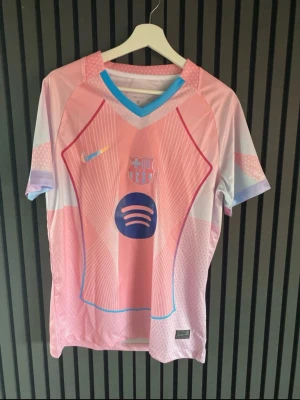 Rosa FC Barcelona matchtröja Nike - Säljer en rosa och ljusblå FC Barcelona matchtröja från Nike med klubbmärke och Spotify-logga på bröstet. Tröjan har korta ärmar, V-ringning med blå kant och sportig design i polyester. Perfekt för fotbollsträning eller att bära som supporter.