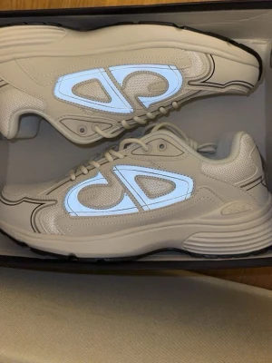 Vita Dior sneakers med reflexdetaljer - Snygga vita sneakers från Dior med grå och reflekterande detaljer på sidorna. Skorna har en chunky sula, meshpaneler och tydliga Dior-loggor. Perfekta för dig som gillar stilrena och moderna sneakers med en twist.