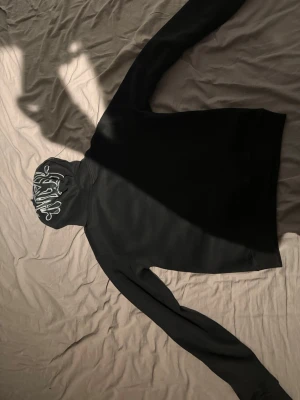 Svart hoodie med graffiti-tryck - Svart hoodie med stor huva och unikt graffiti-inspirerat tryck i vitt på huvan. Hoodien har en klassisk passform och är tillverkad i mjukt material, perfekt för en avslappnad streetwear-look. Det är en syna world hoodie 
