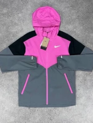 Nike vindjacka rosa och grå - Snygg vindjacka från Nike i rosa, grått och svart. Jackan har huva, dragkedja framtill och två fickor med rosa detaljer. Ikonisk Nike-logga på bröstet. Perfekt för en sportig och trendig look.