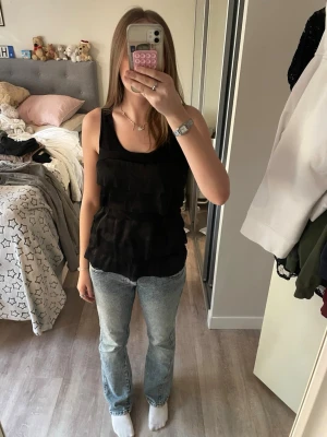 Svart linnetopp med volanger - Svart ärmlös linnetopp med flera lager volanger framtill. Toppen har rund halsringning och är tillverkad i ett mjukt, lätt material som känns skönt mot huden. Perfekt att styla med jeans eller kjol för en trendig look.