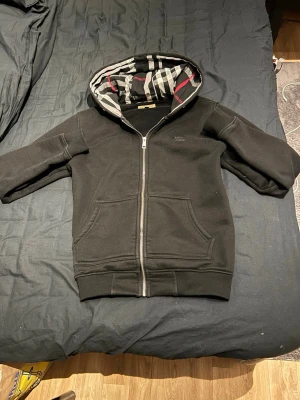 Svart hoodie från Burberry - Svart hoodie med dragkedja från Burberry. Klassisk modell med två fickor fram och huva fodrad i Burberrys ikoniska rutmönster i svart, vitt och rött. Perfekt för en stilren och avslappnad look. Mjuk bomullskvalitet och ribbade muddar.