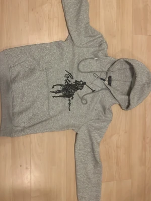 Grå hoodie från Polo Ralph Lauren - Snygg grå hoodie från Polo Ralph Lauren med stor svart logga och polospelare på bröstet. Klassisk känguruficka framtill, justerbar dragsko i huvan och mjukt material som känns skönt mot huden. Perfekt för en avslappnad och trendig look.