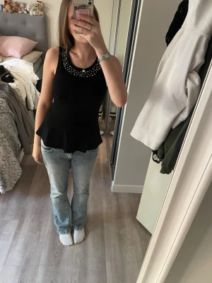 Svart peplumtop med silverdetaljer - Snygg svart ärmlös peplumtop med silvriga nitar runt halsringningen. Toppen har en figurnära passform upptill och utsvängd nederdel. Perfekt att styla med jeans eller kjol för en trendig look.