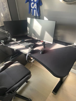Svart hörn-datorbord med skärmfäste - Säljer ett svart hörnformat datorbord med stort musmatteunderlag och dubbla skärmfästen. Perfekt för gaming eller hemmakontor. Bordet har plats för flera skärmar och tillbehör, och har en modern, stilren design i metall och tyg.