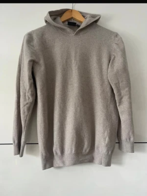 Kashmir hoodie Oscar Jacobsson - En stilren beige hoodie med lång ärm och ribbade muddar. Hoodien har en enkel design utan tryck eller fickor och är tillverkad i ett mjukt, stickat material som känns skönt mot huden. Perfekt för dig som gillar en clean och avslappnad look.