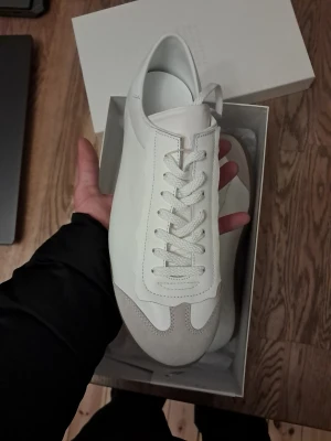 Vita sneakers från Maison Margiela - Stilrena vita sneakers från Maison Margiela med klassisk låg siluett. Skorna har ovandel i vitt läder med detaljer i ljusgrå mocka och vit sula i gummi. Perfekta för dig som gillar minimalistisk och clean stil.