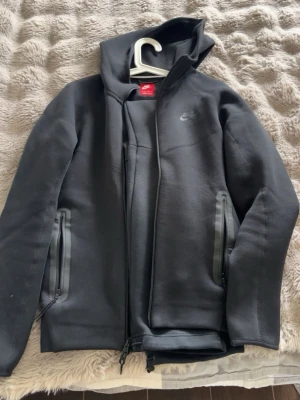 Nike Tech Fleece set i svart - Nike Tech Fleece set med hoodie och byxor i svart färg. Hoodien har dragkedja, huva och två fickor med dragkedja. Byxorna har dragkedjefickor. Båda plaggen har diskret Nike-logga. Perfekt för en sportig och avslappnad stil.