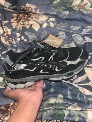 Svarta och silver Asics sneakers - Snygga sneakers från Asics i svart och grå med coola detaljer och meshpaneler för extra andningsförmåga. Skorna har en chunky sula och klassisk snörning. Perfekta för dig som gillar sportig stil och vill sticka ut med ett par trendiga sneakers.