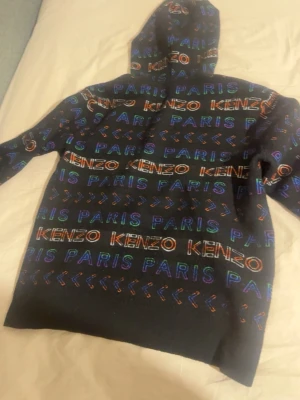 Svart Kenzo hoodie med färgglatt tryck - Cool svart hoodie från Kenzo med färgglada broderade loggor och texten 'PARIS' över hela tröjan. Hoodien har en klassisk passform med huva och långa ärmar. Perfekt för dig som vill sticka ut med en streetstyle-look.
